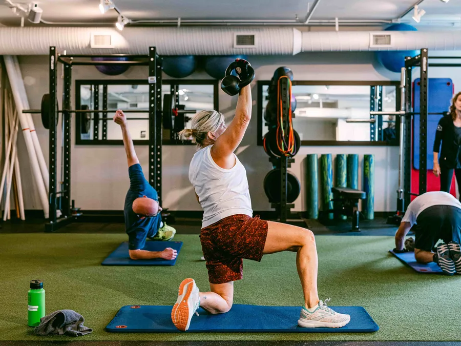 A member in a split lunge pressing a kettlebell overhead on the green turf lane while a coach watches.