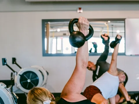 A member braced on one hand and knee, reaching a kettlebell overhead, rowers behind.
