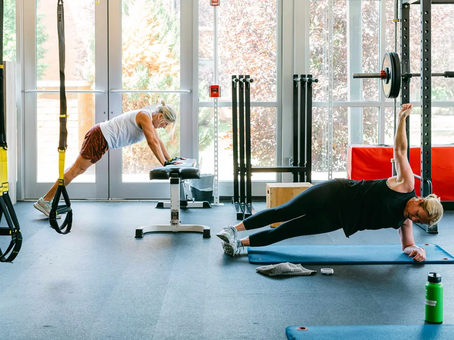 A member in a side plank with one hand on a bench and the other arm raised, TRX straps and windows behind.