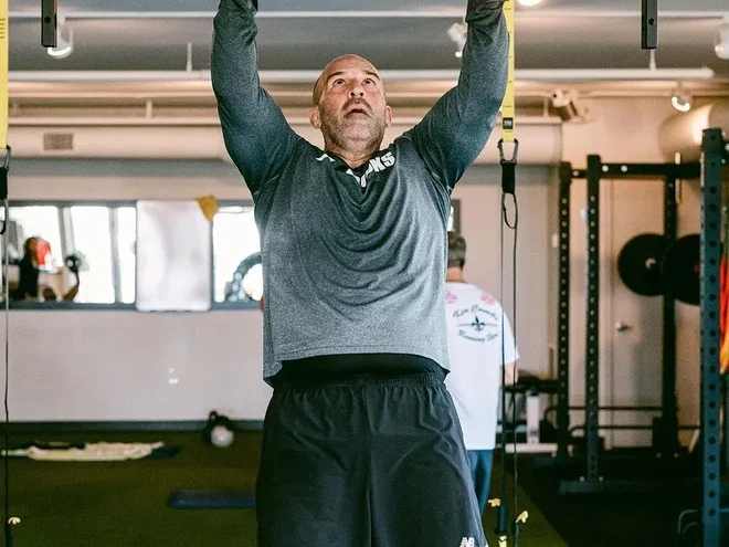 A member reaching both arms overhead under a coach’s eye, with the racks and rowers behind.