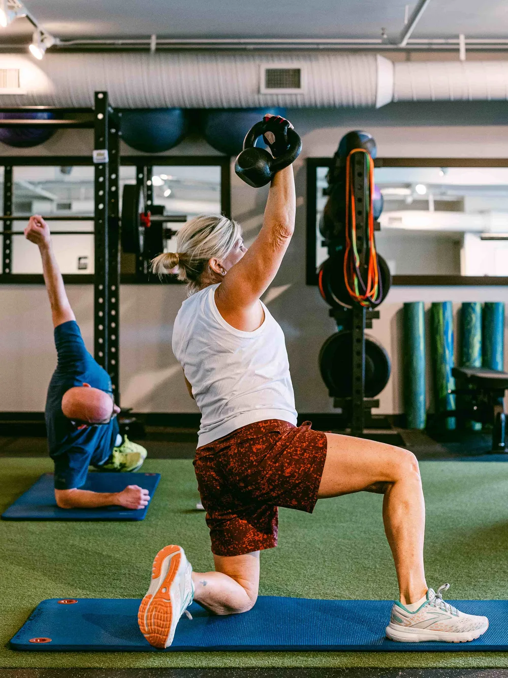 A member in a split lunge pressing a kettlebell overhead on the turf lane during a coached group session.