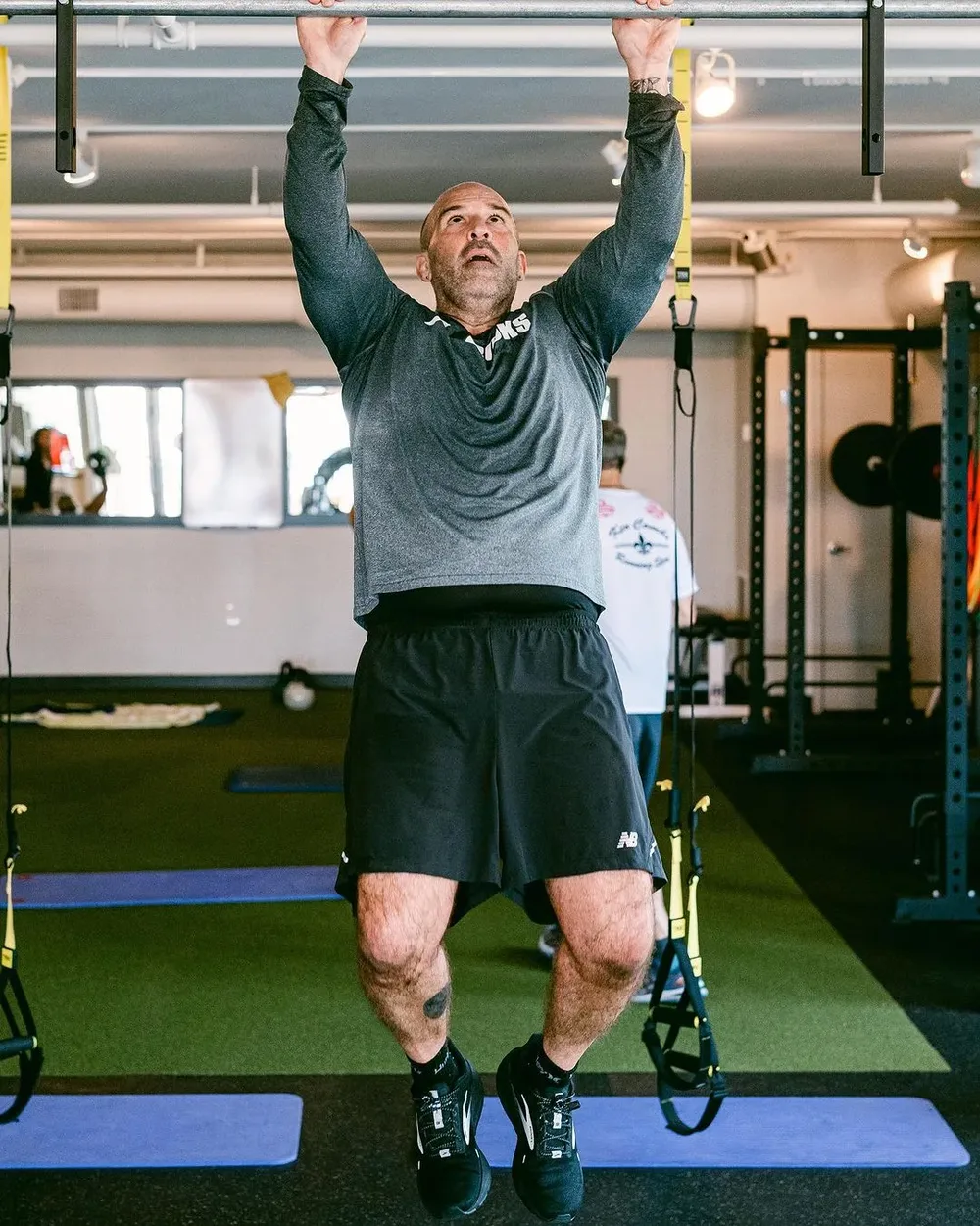 A member hanging from the pull-up bar at the top of a rep on the turf lane, TRX straps either side and the racks behind.