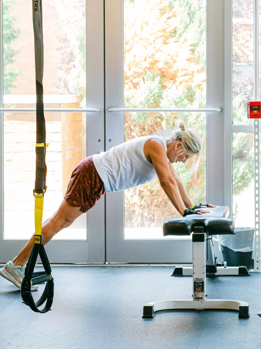 A member holding an incline push-up on a bench in front of the studio's glass doors, TRX straps beside her.